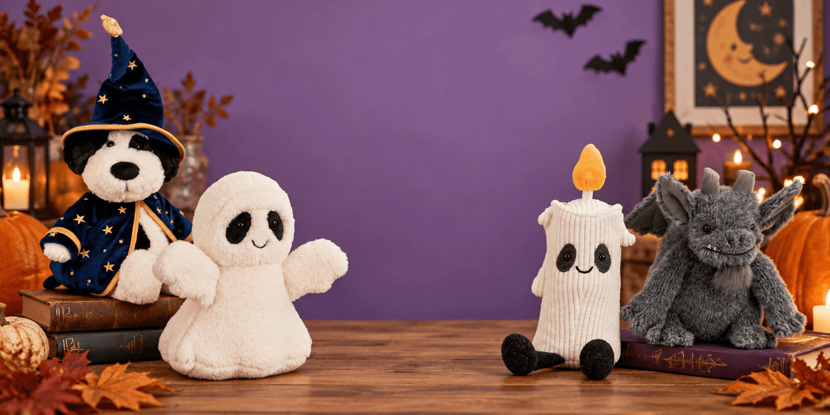A Halloween-themed plush toy display on a wooden table against a purple wall. The collection includes a white ghost plush, a white candle-shaped plush with a yellow flame, a gray dragon plush, a black-and-white dog plush wearing a wizard hat, and a small pumpkin plush.