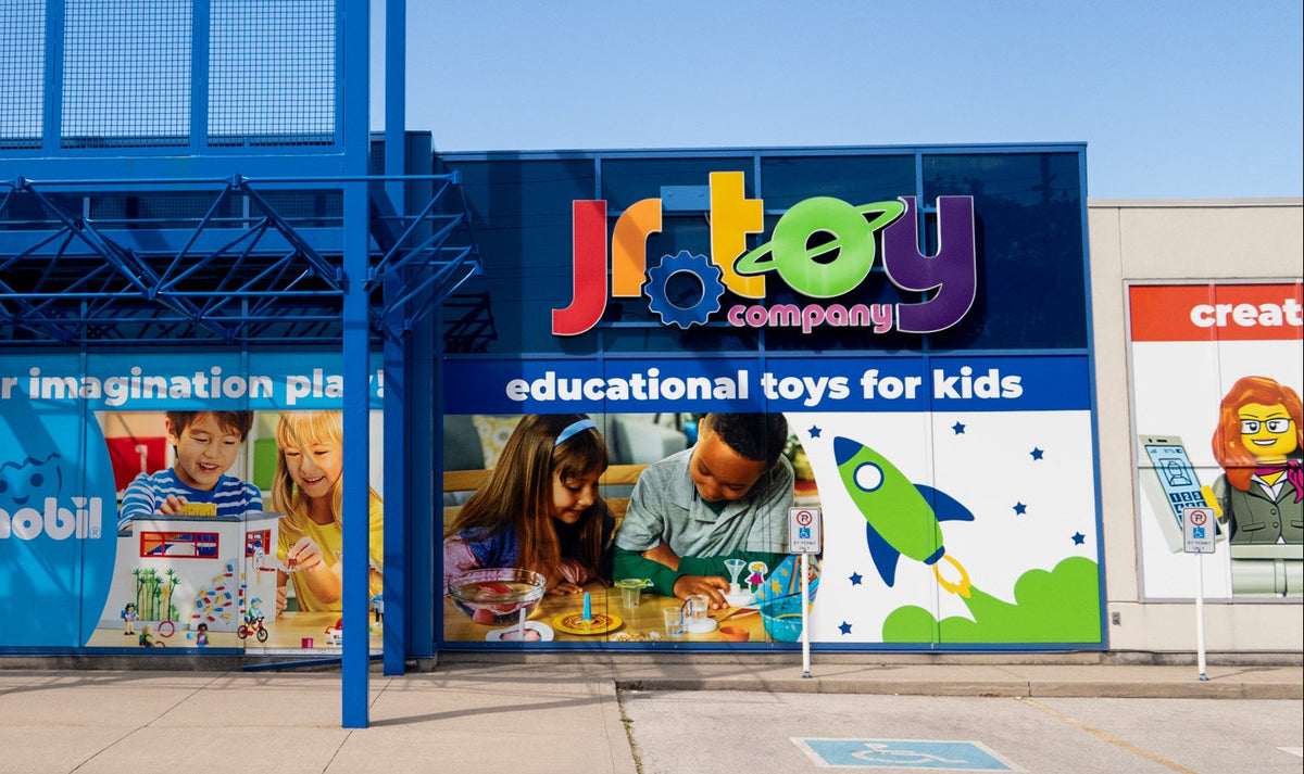 Jr. Toy Company storefront with blue signage and large colorful promotional panels advertising educational toys for kids, photographed outdoors under a clear blue sky.
